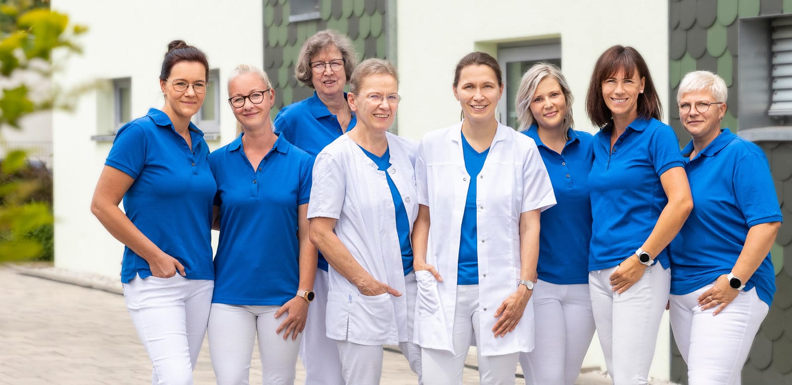 Acht Frauen in weißer Hose und blauem Shirt vor dem grünen Praxisgebäude stehend